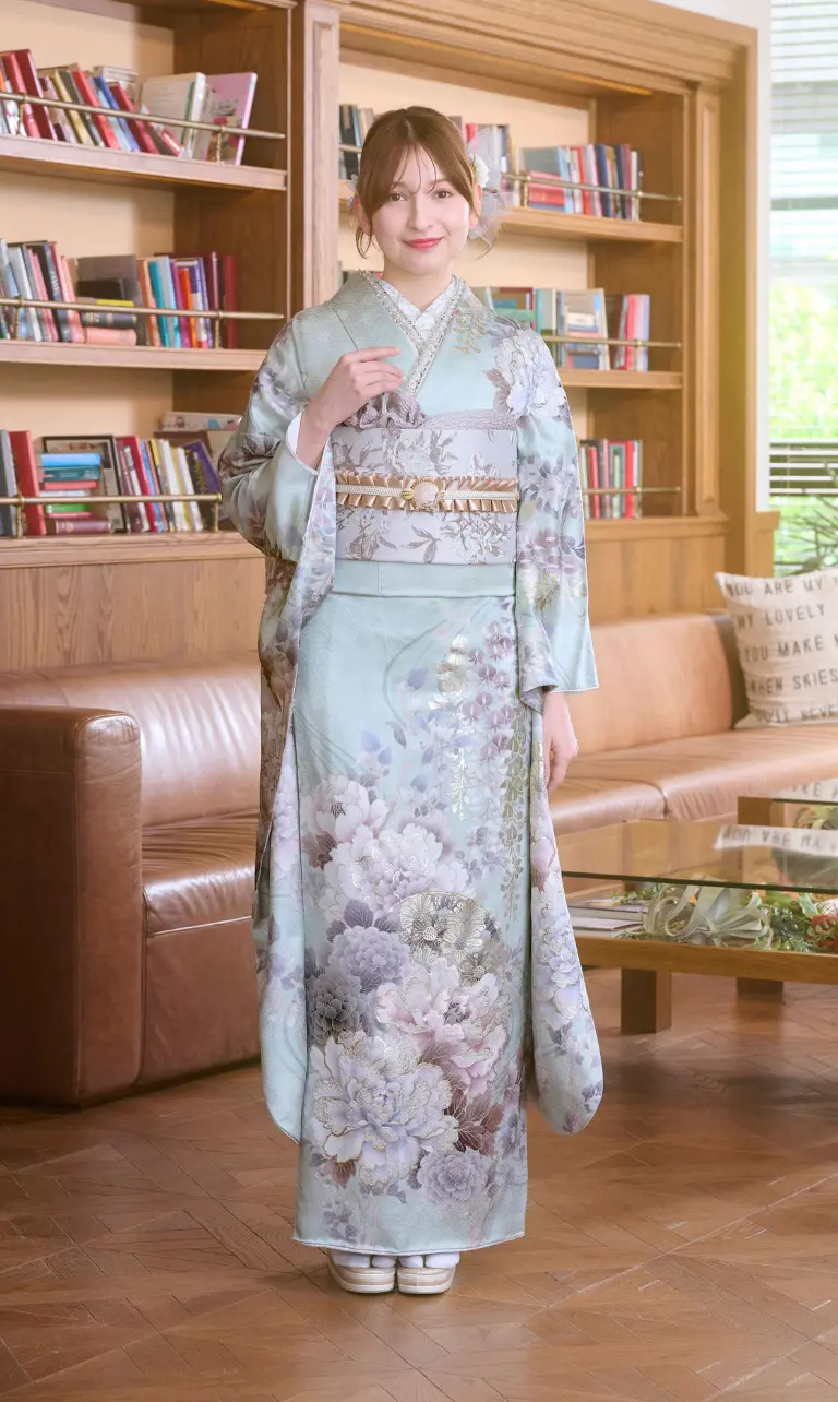 M-501の淡いブルーの花柄着物を着た女性が、屋内に立っている。彼女は本棚と茶色の革張りのソファの前におり、木製の床とガラスのコーヒーテーブルが置かれた、図書室や読書室のような空間にいる。.