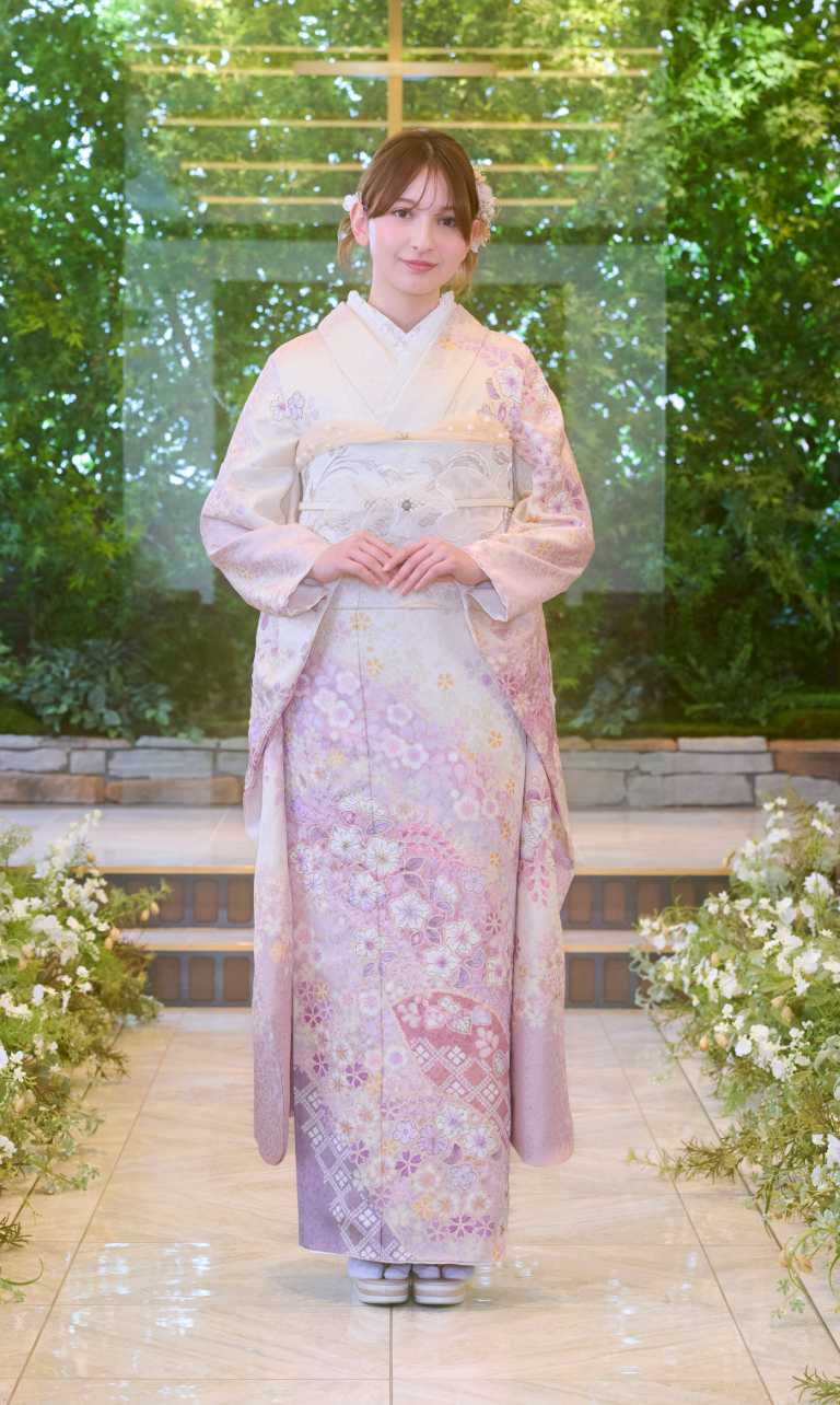 ある女性が、淡いピンクの花柄の着物「M-502」を身にまとい、伝統的な草履を履いて、庭園のような場所で立っている。髪にはピンクの花がきれいに飾られており、両手を胸の前で組んで立っている。.