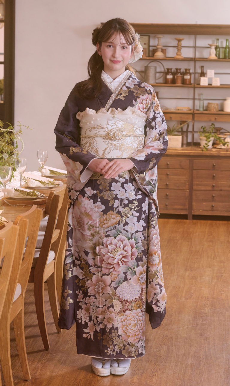 室内で、M-504の伝統的な濃い色の花柄の着物に、淡い色の帯、白い足袋、草履を履いた女性が立っている。彼女は両手を胸の前で組んでほほえんでおり、背景には木製の棚とダイニングテーブルが見える。.