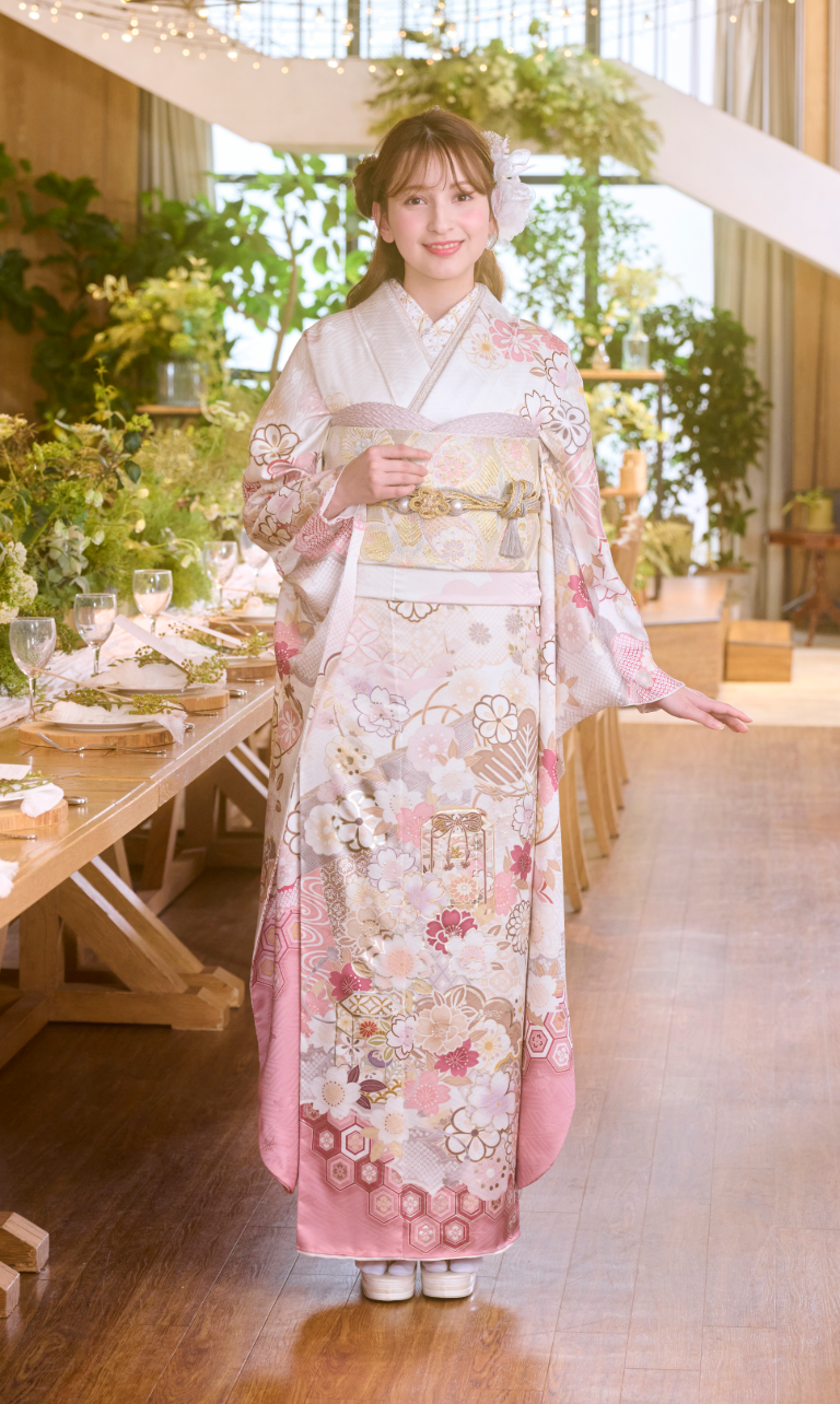 室内で、M-517のピンクと白の花柄の着物を着た女性が、片手を上げて微笑んでいる。華やかに飾られた食卓と、青々とした観葉植物が背景を明るく彩っている。.