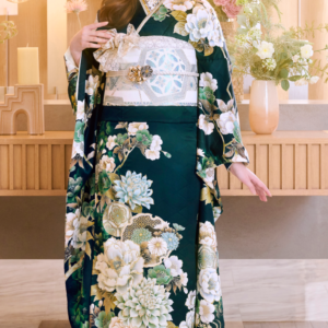 ある女性が、装飾的な帯を締めた濃い緑色の花柄の着物「M-506」を身にまとい、屋内に立っている。彼女は着物の裾を手に持ちながらうつむいており、背景には花々や柔らかな照明が映り込み、優雅で穏やかな雰囲気を醸し出している。.