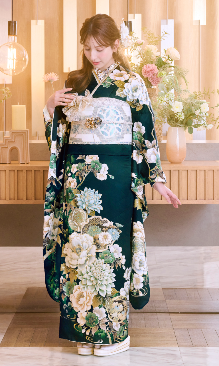 ある女性が、装飾的な帯を締めた濃い緑色の花柄の着物「M-506」を身にまとい、屋内に立っている。彼女は着物の裾を手に持ちながらうつむいており、背景には花々や柔らかな照明が映り込み、優雅で穏やかな雰囲気を醸し出している。.