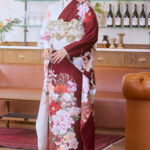 M-506の赤と白の花柄の着物を着た女性が、木製の床の上で室内に立っており、髪には花を飾っている。彼女の背後の棚には、瓶やグラス、鉢植えが並べられている。.