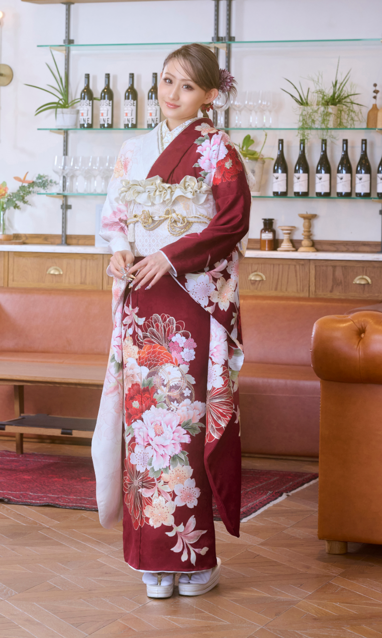 M-506の赤と白の花柄の着物を着た女性が、木製の床の上で室内に立っており、髪には花を飾っている。彼女の背後の棚には、瓶やグラス、鉢植えが並べられている。.