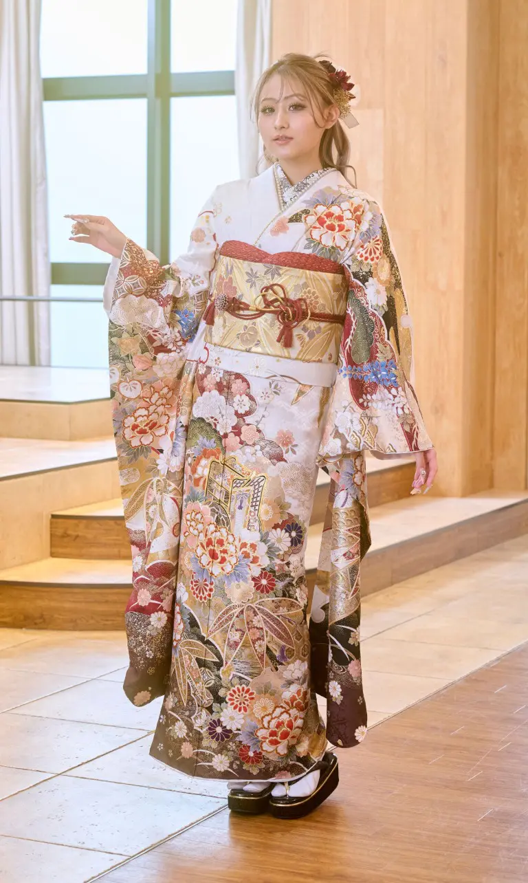 室内で、金、赤、白の鮮やかな花柄が特徴の「M-501」という着物を着た人物が立っています。白い足袋と黒い草履を合わせています。髪は花飾りを付けてまとめられており、右手で指さしています。.