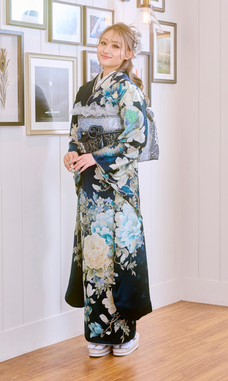 室内で、伝統的な紺色の花柄の着物「M-517」を身にまとい、白いサンダルを履いた人物が立っている。背景には額に入った写真や白いパネルの壁が見え、床は木製で、頭上にはペンダントライトが吊り下げられている。.