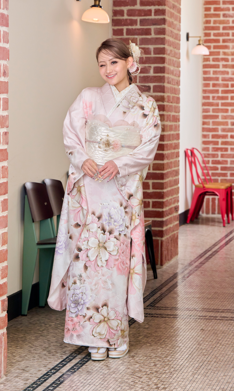 淡いピンクの花柄の着物「M-511」を着た人物が、屋内のレンガの壁のそばで、背景に赤と緑の椅子や壁灯を背に、笑顔でポーズをとっている。.