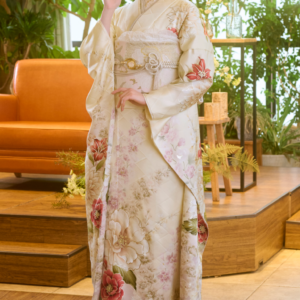 室内で、ピンクの花柄をあしらったクリーム色のM-506の着物を着た女性が、片手を顔のそばに当てて立っている。髪には花を挿しており、周囲には植物や木製の家具が置かれている。.