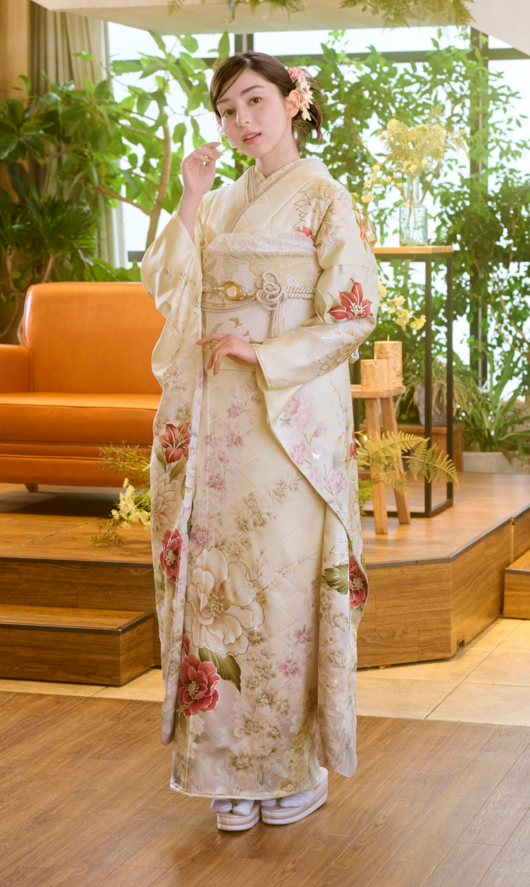 室内で、ピンクの花柄をあしらったクリーム色のM-506の着物を着た女性が、片手を顔のそばに当てて立っている。髪には花を挿しており、周囲には植物や木製の家具が置かれている。.
