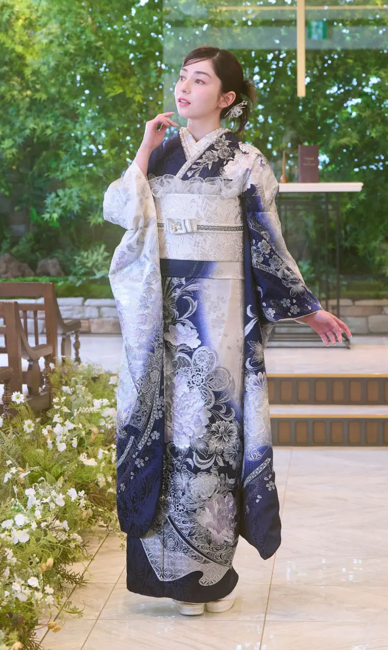 ある女性が、室内で花のそばに立っている。彼女はネイビーと白の花柄の「M-501」着物を着ている。彼女は横を向き、左手を顔の近くに当てている。背後の窓からは緑が見えている。.
