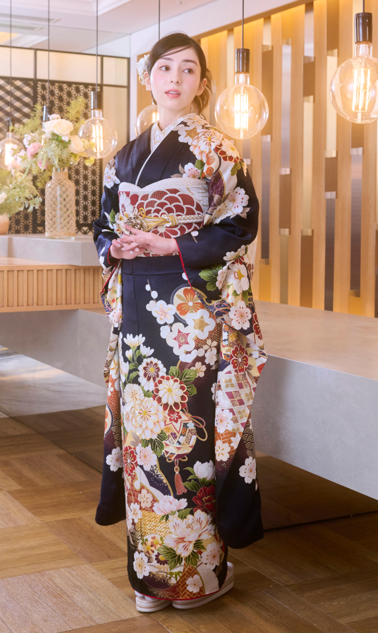 ある女性が、精巧な模様が施された濃い色の花柄の着物「M-517」を身にまとい、屋内に立っている。彼女は両手を合わせ、吊り下げられた電球や生け花に囲まれ、モダンで優雅な空間の中に佇んでいる。.