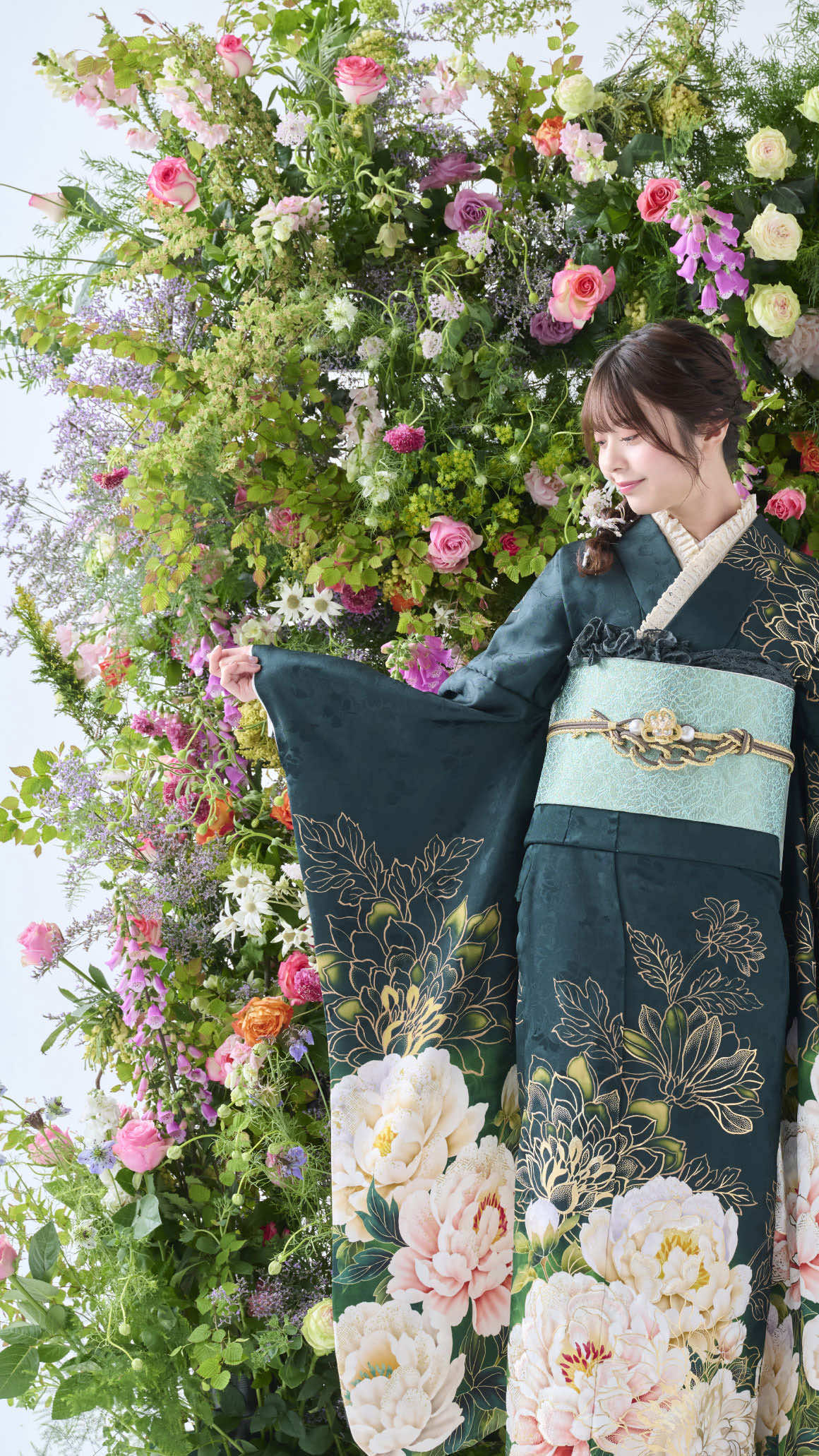 濃い色の花柄の着物を着た人物が、バラや様々な緑が描かれた鮮やかな花の背景の前に立っています。着物には繊細な花模様が施されており、人物は袖口を優しく押さえながら下を向いています。.